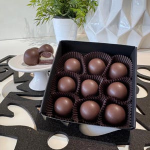 May include: A box of nine chocolate truffles, arranged in a grid pattern. The truffles are dark brown and have a smooth, glossy finish. The box is black and has a white paper liner.