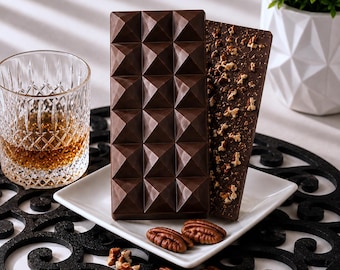 May include: Two rectangular dark chocolate bars on a white square plate. One bar features a geometric pyramid-shaped mold, while the other is topped with chopped pecans. Several whole pecans are scattered on the plate and surrounding surface.