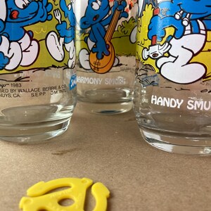 Pu&ograve; includere: Tre bicchieri da bere in vetro vintage con personaggi dei Puffi. I bicchieri sono trasparenti con illustrazioni di cartoni animati dei Puffi in blu e giallo. Il testo sui bicchieri recita "Harmony Smurf" e "Handy Smurf".