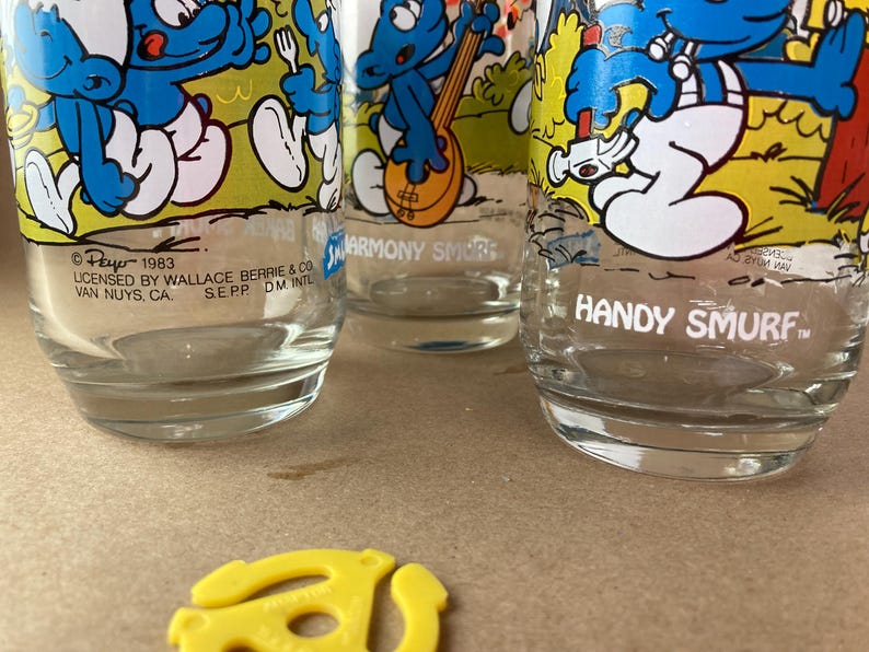 Pu&ograve; includere: Tre bicchieri da bere in vetro vintage con personaggi dei Puffi. I bicchieri sono trasparenti con illustrazioni di cartoni animati dei Puffi in blu e giallo. Il testo sui bicchieri recita "Harmony Smurf" e "Handy Smurf".