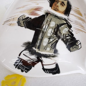 May include: A white ceramic dish with a stylized illustration of a person wearing a black and white patterned jacket and pants. The person is running with their arms outstretched and the background is a blur of gold and brown lines. The dish is signed "Acodin" in the lower left corner.