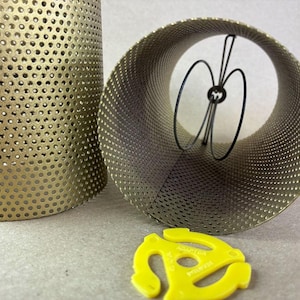 May include: Two perforated metal lamp shades in a gold tone, one upright and the other lying on its side. A yellow 45 RPM record adapter is in the foreground. The shades have a cylindrical shape and are made of a metal mesh.