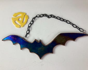 Puede incluir: Un murciélago de vidriera con alas iridiscentes azules y verdes, suspendido por una cadena negra. También hay un adaptador de disco de 45 RPM amarillo. El murciélago es una decoración de Halloween o de temática gótica.