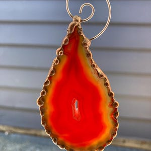 May include: A teardrop-shaped agate slice with a vibrant red centre and a golden-yellow border. The stone is encased in a copper-coloured metal frame with a decorative swirl at the top, suitable for a pendant or ornament.