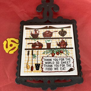 May include: A decorative trivet with a black metal frame and handle. The centre features a ceramic tile with illustrations of kitchenware and the text "THANK YOU FOR THE WORLD SO SWEET THANK YOU FOR THE FOOD WE EAT."