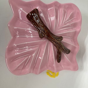 May include: A pink ceramic leaf-shaped dish with a brown twig-shaped accent. The dish is perfect for serving small snacks or holding jewelry.