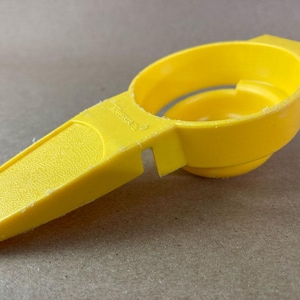 May include: A bright yellow egg separator made of plastic. The separator has a handle with a textured grip and a circular bowl for catching the yolk. The design includes a small hole in the handle. A kitchen utensil for separating egg whites.