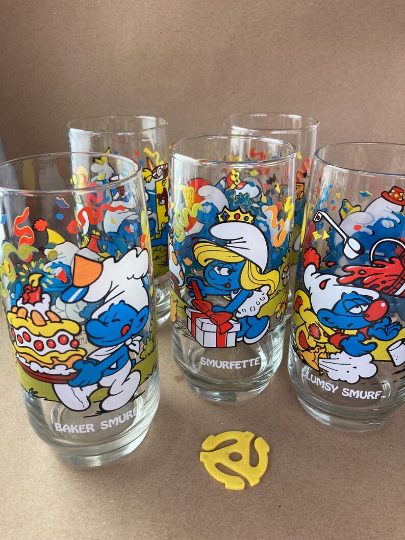 Pu&ograve; includere: Set di quattro bicchieri da bere a tema Puffi. Ogni bicchiere presenta un diverso personaggio dei Puffi con uno sfondo colorato. I bicchieri sono trasparenti e hanno una forma arrotondata. I Puffi sono raffigurati in una scena di festa con coriandoli e palloncini. I bicchieri sono etichettati con i nomi dei Puffi: Puffo Panettiere, Puffetta, Puffo Sbadato.
