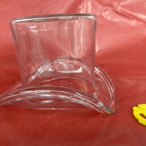 Peut inclure: Un chapeau en verre transparent en forme de chapeau haut de forme avec un large bord. Le chapeau est posé sur une surface rouge.