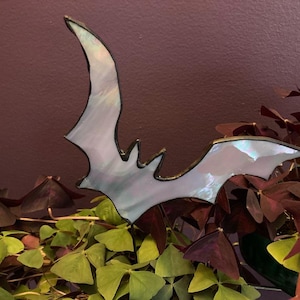 May include: A stained glass bat decoration with iridescent wings and a black outline. The bat is set against a background of green and burgundy foliage, creating a Halloween or gothic theme.