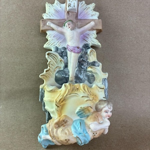 May include: A religious figurine depicting Jesus on a cross, with an angel at the base. The cross is brown with a white and purple halo. The angel is in a kneeling position with blue and tan clothing.