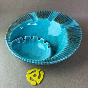 May include: A turquoise ceramic ashtray with a built-in rest and a ridged rim. A yellow 45 RPM record adapter sits below the ashtray. The ashtray has a glossy finish and a few white painted details.