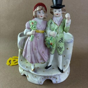 May include: A porcelain figurine featuring a couple in period attire. The woman wears a pink dress and holds a bouquet, while the man in a green coat holds flowers and a top hat. The base is decorated with gold accents.