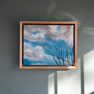 Op de afbeelding: Een ingelijst schilderij van een bewolkte lucht met een silhouet van kale takken op de voorgrond. De lucht is geschilderd in tinten blauw, wit en grijs. De takken zijn zwart.