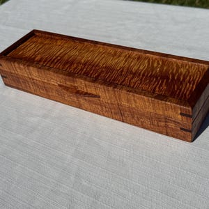 May include: A rectangular wooden box with a lid. The box is made of a light brown wood with a wavy grain pattern. The box is approximately 10 inches long and 4 inches wide.