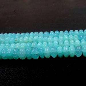 May include: Strands of light blue gemstone beads. Each bead has a rounded, barrel shape with a subtle, carved design. The beads are strung together, creating a continuous line of color and texture. The background is a dark, solid color.