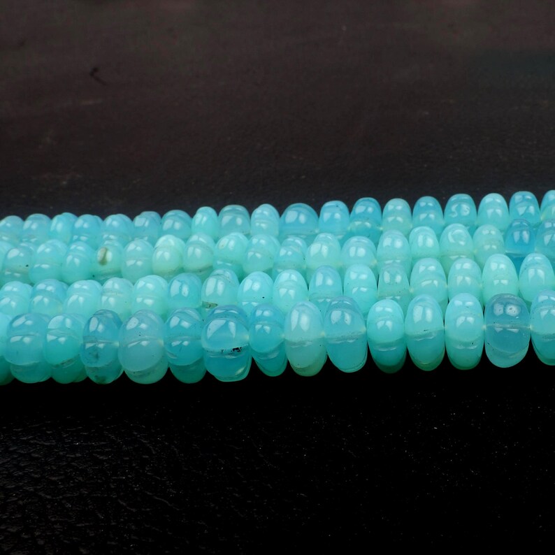 May include: Strands of light blue gemstone beads. Each bead has a rounded, barrel shape with a subtle, carved design. The beads are strung together, creating a continuous line of color and texture. The background is a dark, solid color.