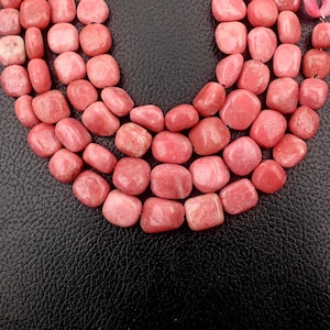 May include: Close-up of a necklace made of pink, rounded gemstone beads. The beads are a mix of light and dark pink hues, strung together. The background is a textured black surface.