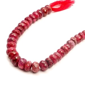 May include: A strand of faceted, round, deep pinkish-red gemstone beads arranged in a curved line. The beads feature a textured, multi-faceted surface that reflects light. A bright red tassel is attached to one end of the string of beads.