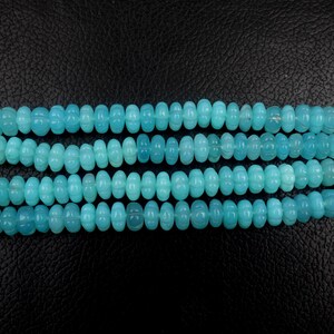 May include: Four strands of light blue gemstone beads. Each bead is oval-shaped with a carved floral design. The beads are strung together, creating a necklace or bracelet component. The background is black.