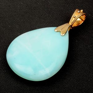 Puede incluir: Colgante en forma de lágrima con una piedra preciosa azul claro. La piedra está montada en un engaste dorado. El colgante se muestra sobre un fondo negro.