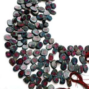 May include: A collection of loose, tumbled gemstone beads featuring a dark teal and blue-grey exterior with vibrant magenta and deep pink centers. The stones are irregularly shaped, flat, and polished, arranged in a curved pattern with red threads.
