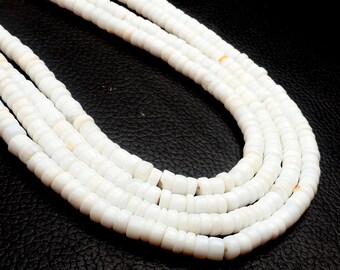 May include: Close-up of multiple strands of white, square-cut beads. The beads are strung together, creating a layered effect. The beads have a slightly off-white color and a smooth texture. The background is a dark, textured surface.