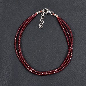 Puede incluir: Una pulsera de tres hilos con pequeñas cuentas facetadas de color rojo intenso. La pulsera tiene un cierre plateado y una cadena ajustable. Las cuentas están ensartadas en un fino cordón rojo oscuro. La pulsera se muestra sobre una superficie oscura.