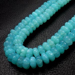 May include: Close-up of three strands of light blue gemstone beads. The beads have a carved, ridged design and are strung together. The background is a dark, textured surface. The beads are a soft, opaque color.