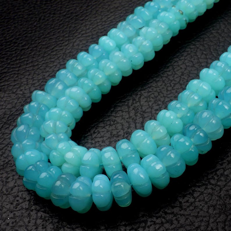 May include: Close-up of three strands of light blue gemstone beads. The beads have a carved, ridged design and are strung together. The background is a dark, textured surface. The beads are a soft, opaque color.