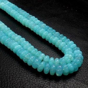 May include: Close-up of three strands of light blue gemstone beads. The beads have a rounded, segmented shape and a translucent quality. The beads are arranged on a dark, textured surface.