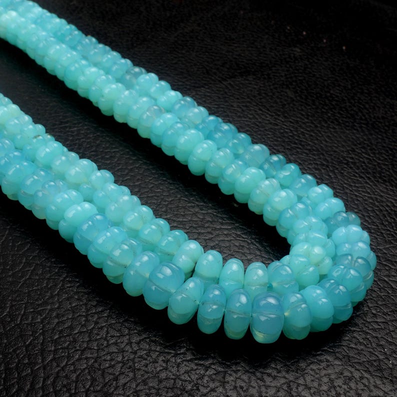 May include: Close-up of three strands of light blue gemstone beads. The beads have a rounded, segmented shape and a translucent quality. The beads are arranged on a dark, textured surface.