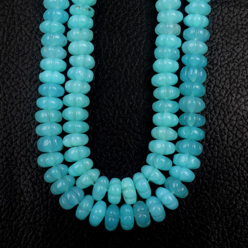 May include: A close-up shot of a necklace made of light blue, carved beads. The beads are oval-shaped with a floral design and are strung together in three strands. The background is a dark, textured surface.