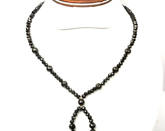 May include: A black beaded necklace with a teardrop pendant. The necklace is made of small, faceted beads and larger, round beads. The teardrop pendant is made of graduated beads. The necklace is displayed on a white mannequin.