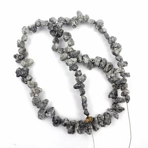 Puede incluir: Un hilo de diamantes en bruto, sin tallar, de color gris y negro, ensartados en un alambre fino. Las piedras son de formas y tamaños irregulares, con una superficie rugosa y texturizada de brillo metálico. Se observa una pequeña piedra dorada.