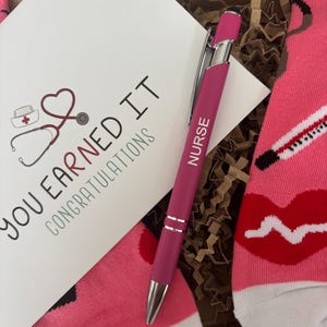 May include: A pink and white pair of socks with a red lip, a stethoscope, and a thermometer design. A pink pen with the word "NURSE" printed on it is included. A card with the text "YOU EARNED IT CONGRATULATIONS" is also included.