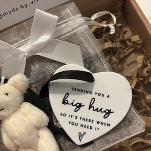 以下が含まれることがあります： 白いセラミック製のハート型のオーナメントで、黒い文字で「Sending you a big hug so it's there when you need it」と小さなハートのシンボルが書かれています。オーナメントは白いオーガンザの袋に入っていて、白いテディベアが入っています。