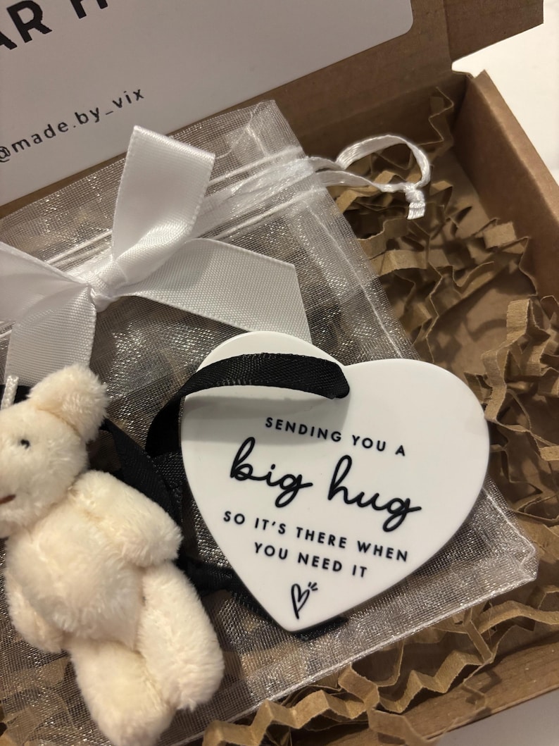 以下が含まれることがあります： 白いセラミック製のハート型のオーナメントで、黒い文字で「Sending you a big hug so it's there when you need it」と小さなハートのシンボルが書かれています。オーナメントは白いオーガンザの袋に入っていて、白いテディベアが入っています。