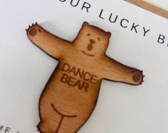 May include: A wooden cutout of a bear with outstretched arms, the bear is brown and has the words "Dance Bear" written on its chest. The bear is on a white card with the text "I am your lucky bear" and "Take me in your dance bag, take me everywhere".
