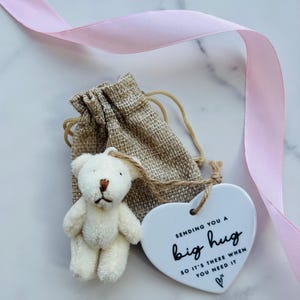 May include: A small cream-colored plush teddy bear attached to a white heart-shaped ceramic ornament with the black text "SENDING YOU A big hug SO IT'S THERE WHEN YOU NEED IT" and a small heart icon, all placed in a tan burlap drawstring pouch.