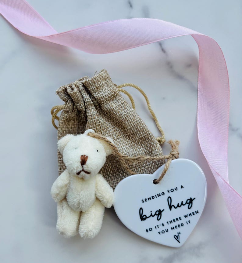 May include: A small cream-colored plush teddy bear attached to a white heart-shaped ceramic ornament with the black text "SENDING YOU A big hug SO IT'S THERE WHEN YOU NEED IT" and a small heart icon, all placed in a tan burlap drawstring pouch.