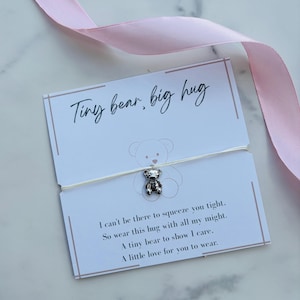 May include: A silver-toned bear charm on a thin white string, attached to a white card with the text Tiny bear, big hug and a poem. The card features a faint bear outline and is accented by a light pink ribbon draped across the top corner.