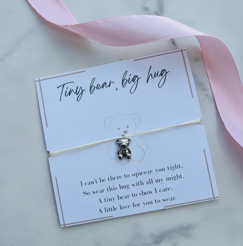 May include: A silver-toned bear charm on a thin white string, attached to a white card with the text Tiny bear, big hug and a poem. The card features a faint bear outline and is accented by a light pink ribbon draped across the top corner.