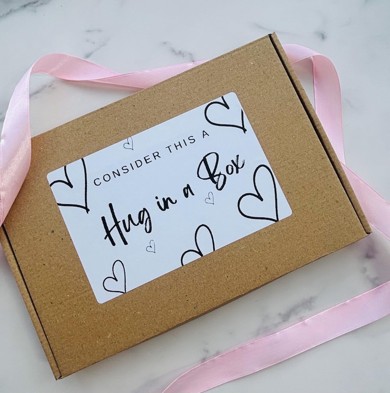 May include: A rectangular brown cardboard box featuring a white rectangular label with black text that reads "CONSIDER THIS A Hug in a Box" surrounded by hand-drawn black heart outlines. A light pink satin ribbon is wrapped around the box.