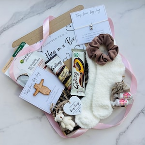 May include: A 'Hug in a Box' gift set containing a face mask, Galaxy chocolate, hot chocolate sachet, fluffy socks, a scrunchie, a teddy bear, and a bracelet. The box is decorated with a pink ribbon and includes a card.