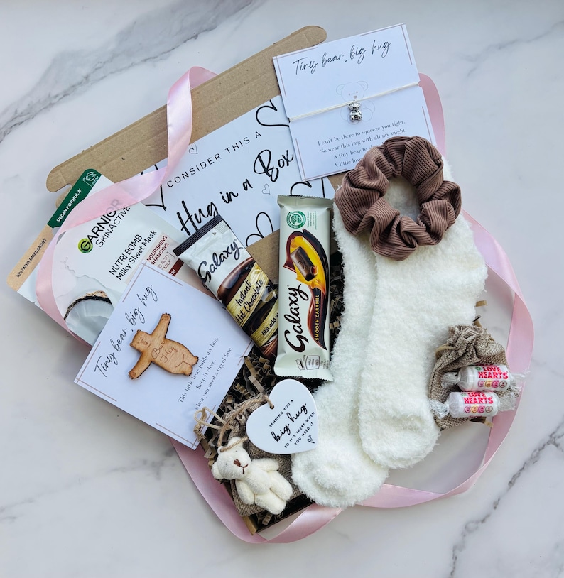 May include: A gift box labeled "Consider This a Hug in a Box" containing a Garnier sheet mask, Galaxy hot chocolate, a Galaxy chocolate bar, a brown scrunchie, white fuzzy socks, two Love Hearts candies, a small teddy bear, and two cards.