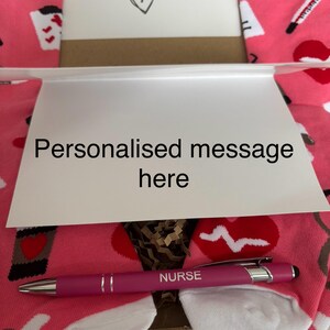 May include: A pink and white pair of socks with a stethoscope, a thermometer, and a heart beat graphic. A pink pen with the word "NURSE" engraved on it. A card with the text "YAY! YOU QUALIFIED" and a heart. A white card with the text "Personalised message here".