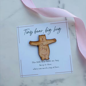 May include: A light brown wooden bear cutout with arms outstretched, featuring the engraved text Bear Hug. It is mounted on a white card with the printed text Tiny bear, big hug and This little bear holds my hug. Keep it close, when you need a tug of love.
