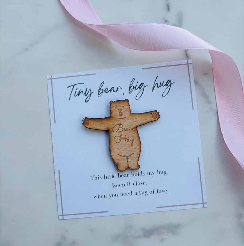 May include: A light brown wooden bear cutout with arms outstretched, featuring the engraved text Bear Hug. It is mounted on a white card with the printed text Tiny bear, big hug and This little bear holds my hug. Keep it close, when you need a tug of love.