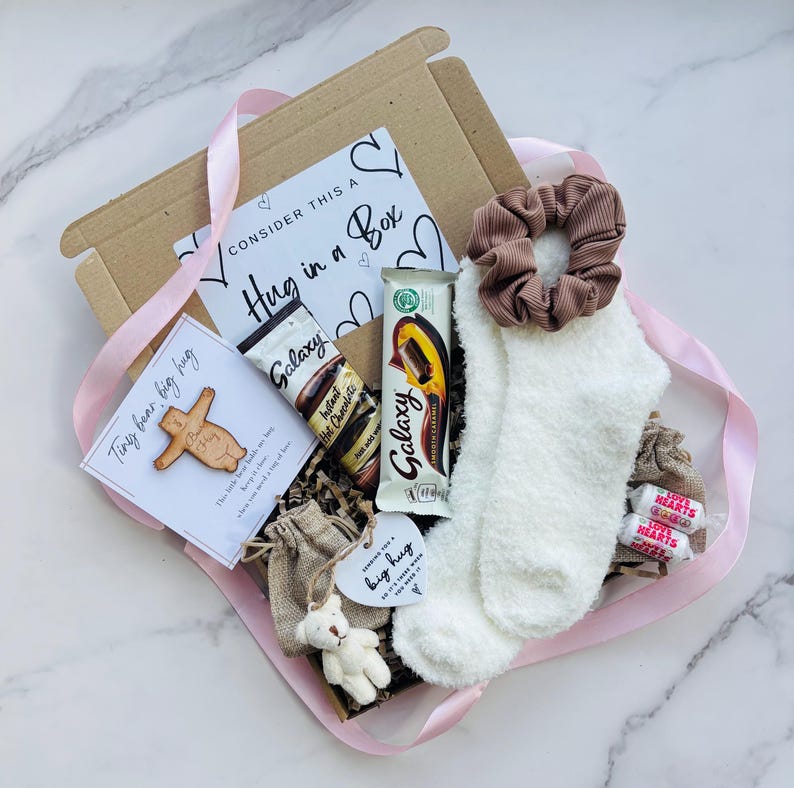 May include: A gift box containing a pair of white fuzzy socks with a brown ribbed scrunchie, a Galaxy hot chocolate sachet, a Galaxy Smooth Caramel chocolate bar, two Love Hearts candies, a small bear figurine, and cards reading Hug in a Box.